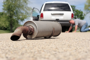 There is a muffler laying in the road in the foreground. In the background a man is looking under his van to see what happened.